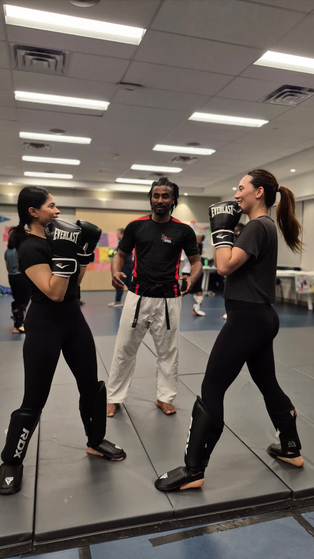 Two adult students in gloves and shin guards face off as the instructor coaches between them