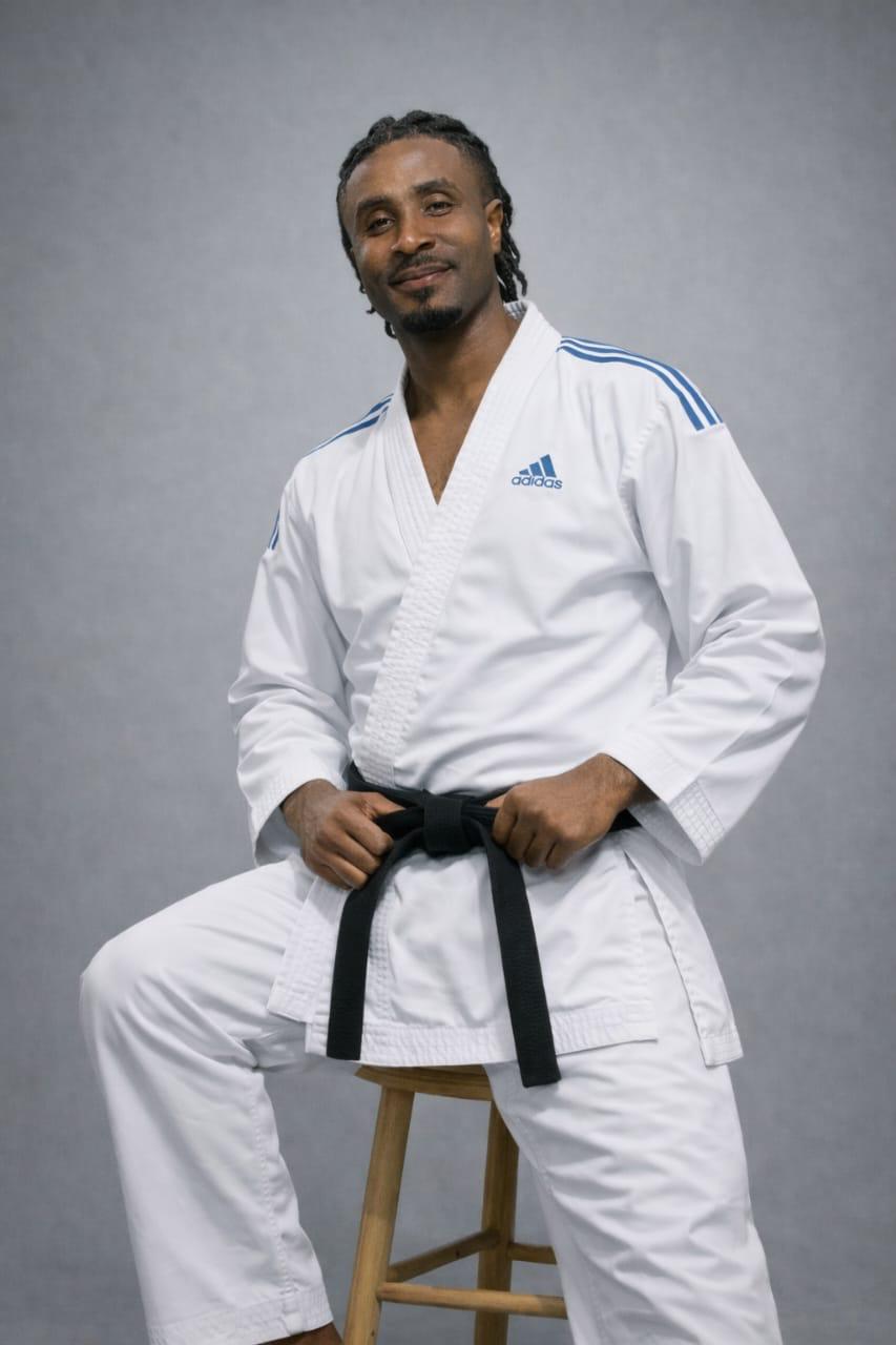 Founder Ifeanyi Ogene seated in a white adidas gi with a black belt, gentle smile, neutral studio background.